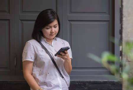 Asian sad woman checking smartphone online content while standing near vintage wall in public placeの写真素材
