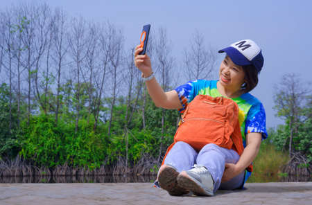 Asian female tourist in colorful summer style smiling cheerfully while using smartphone taking selfie with leafless forest on blue sky at viewpoint in coastal natural parklandの写真素材