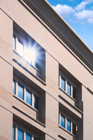 Sunlight reflection on surface of multiple blue glass windows on gray office building wall against white cloud and blue sky in vertical frameの写真素材