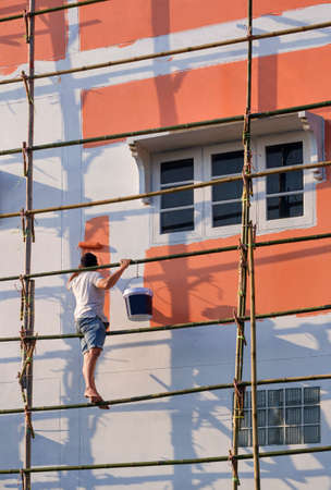 Low angle view of Asian builder worker on wooden scaffolding is painting wall outside of the old house in vertical frameの写真素材