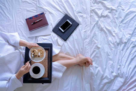 Top view of woman in white bathrobe drinking morning coffee with breakfast after working online on the bedの写真素材