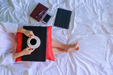 Top view of woman in white bathrobe drinking morning coffee while relaxing after working online on her bedの写真素材