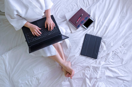 Top view of woman in white bathrobe using laptop computer with book and various device on bed in her bedroomの写真素材
