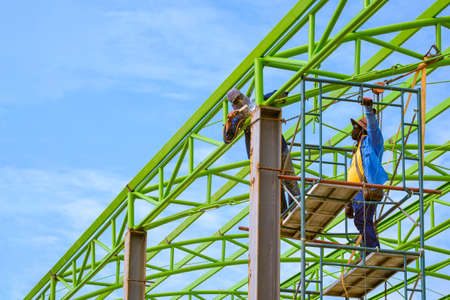 Asian foreman and construction worker on scaffolding are welding metal roof structure of industrial building against blue sky in perspective side viewの写真素材
