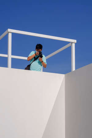 Young Asian man taking picture with digital camera on rooftop of white building against blue clear...