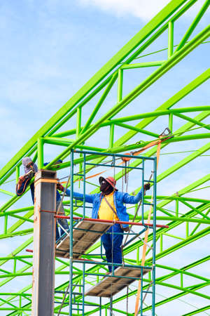 Asian foreman and construction worker on scaffolding are welding metal roof structure of industrial building against blue sky in vertical frameの写真素材