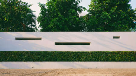 Front view of white concrete wall decoration with bush fence and green trees in front of homeの写真素材