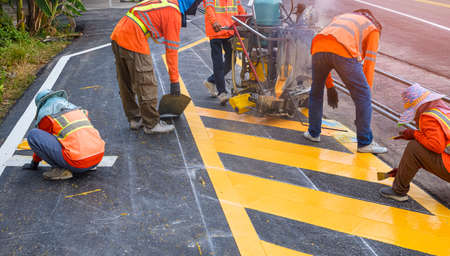 Road workers group with thermoplastic spray marking machine are painting yellow traffic sign on asphalt road with railway track crossing on street surfaceの写真素材