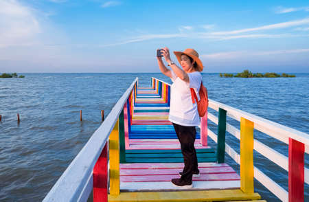 Happy Asian female tourist photographing with smartphone on colorful wooden bridge at sea viewpoint...