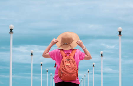 Rear view of female tourist in pink t-shirt holding hat with blurred row of street lamp posts against cloudy sky background at sea viewpoint in pastel color tone, summer and travel conceptの写真素材