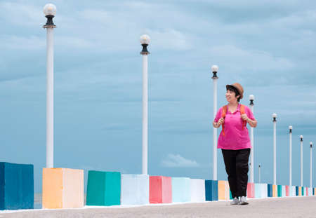 Full length view of happy Asian female tourist walking on multicolored bridge at sea viewpoint against cloudy sky backgroundの写真素材