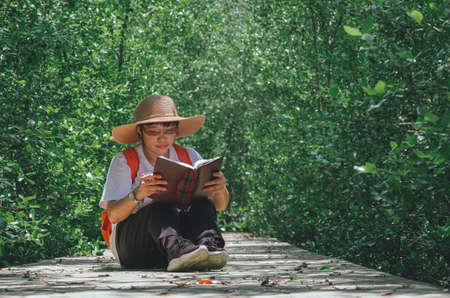 An Asian female forester sitting on walkway and taking note in mangrove forest at natural parkland