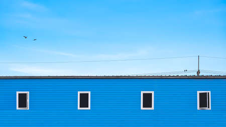 Minimal background of 4 Glass windows on blue wooden wall of row house with birds perching on electric cable lines against blue sky in widescreen viewの写真素材