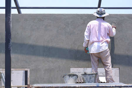 Rear side view of Asian builder worker on scaffolding using wooden trowel polishing cement wall in construction siteの写真素材
