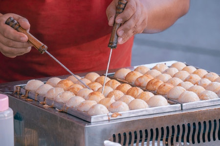 Selective focus at Japanese tasty Takoyaki balls in stainless steel molded stove at outdoor market place with blurred hand while cooking, street food conceptの写真素材