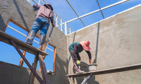 Low angle view of 2 Asian builder workers on wooden scaffolding are plastering concrete wall in house construction siteの写真素材