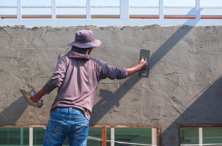 Rear view of builder worker using trowel to plastering cement on the wall inside of house construction siteの写真素材