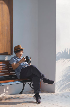 Asian female tourist checking photos on digital camera screen while sitting on bench near white wall outside of vintage building, perspective side view and vertical frameの写真素材