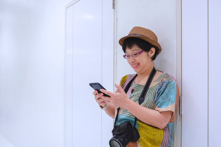 Smiling Asian female tourist in vintage style using smartphone while standing on white wall inside of buildingの写真素材