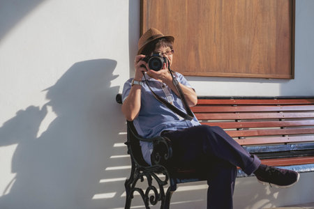 Asian female tourist photographing with digital camera while sitting on bench with white cement wall in vintage tone styleの写真素材