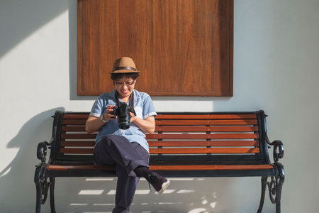 Happy Asian female tourist checking photos on digital camera screen while sitting on bench in front of white vintage building wall, front view with copy spaceの写真素材