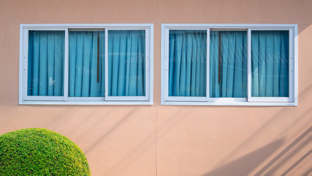 Row of glass sliding windows and blue sunshade on beige gypsum board wall with round shrub on foreground, home exterior architecture conceptの写真素材