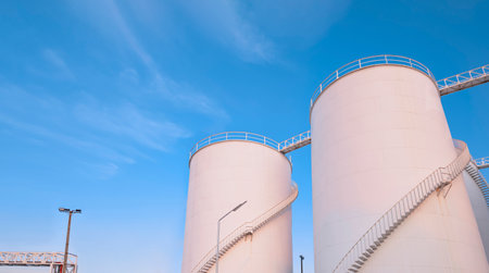 Low angle view of storage fuel tanks with oil pipeline system in petroleum industrial area at harbor against blue sky backgroundの写真素材