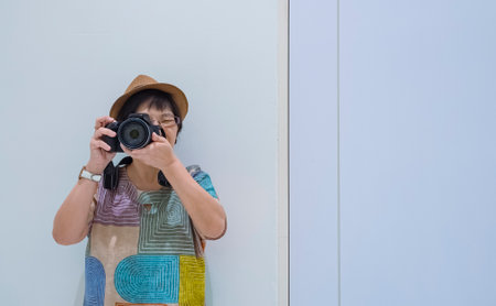 Front view of Asian female tourist photographing with digital camera while leaning against the wall inside of white roomの写真素材