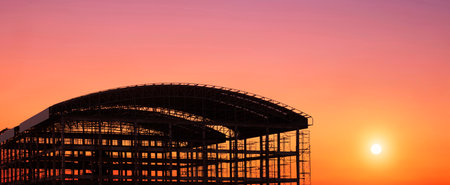 Silhouette of large Industrial Building Structure with curved roof in Construction Site against colorful Sunset sky Background in Panoramic viewの写真素材