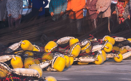Focus at yellow pvc fishing buoys float of black otter trawl on the floor with blurred background of fishermen group are repairing broken netsの写真素材