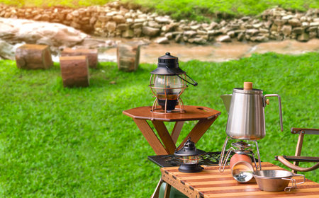 Stainless steel kettle, portable gas stove and vintage lanterns on wooden picnic table set near the stream in natural viewの写真素材