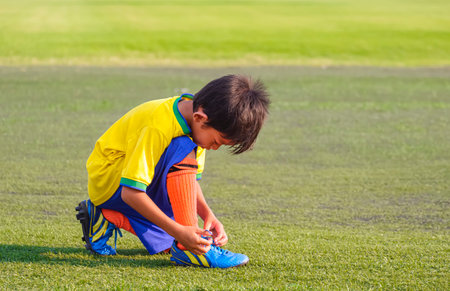 Asian little boy in soccer uniform tying shoelace on soccer field before trainingの写真素材