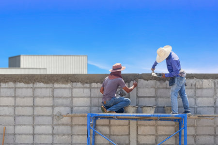 Two Asian Construction Workers on scaffolding are Plastering Cement on edge of concrete Blocks Fence Wall in front of industrial Buildingの写真素材