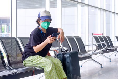 Asian casual female tourist wearing protective mask with suitcase is using smartphone on bench while waiting for boarding in passenger rest area at airport terminalの写真素材