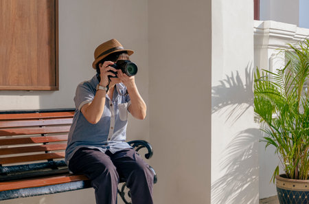 Asian female Tourist Photographing with digital Camera while sitting on bench near white wall outside of Vintage buildingの写真素材