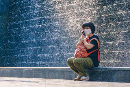 Asian female tourist in protective mask talking on smartphone while sitting on the edge of waterfall stone wall decoration in downtown streetの写真素材