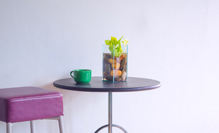 Green coffee mug and houseplant in glass vase on round table with leather chair near empty gray interior wall in minimal styleの写真素材