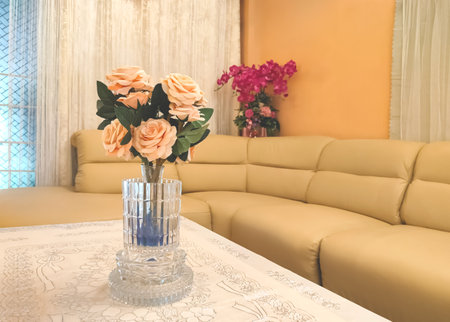 Selective focus at beautiful artificial roses bouquet in crystal vase on a table covered with lace tablecloth with beige sofa in modern living room.の写真素材
