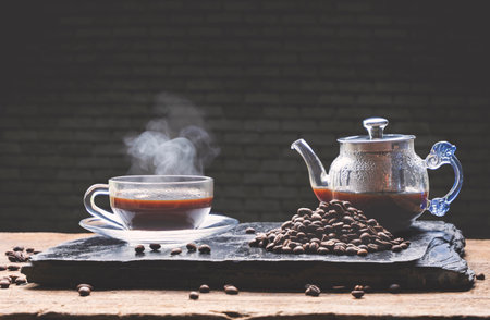 Hot black coffee in glass coffee cup with steam and coffee pot on wooden table with black brick wall...