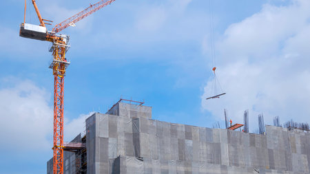 Tower crane is lifting steel bars onto top of high building structure in construction site against blue sky background.の写真素材