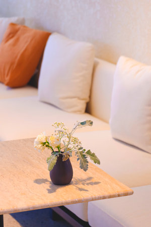 Focus at white flowers in little black terracotta vase on marble desk with blurred cushions on couch in beige tone style and vertical frame, home interior modern loft decoration design conceptの写真素材