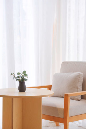 Gray cushion on armchair with ceramic vase on round wooden desk near white curtain inside of modern coffee shop in beige tone style and vertical frameの写真素材