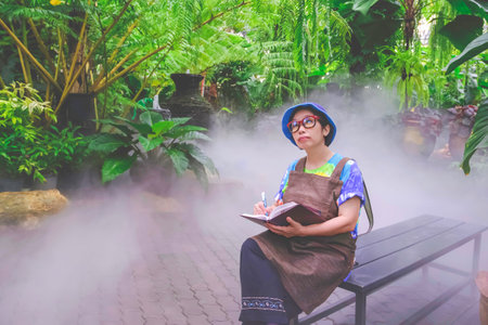 Asian adult woman sitting on bench and taking notes while studying the ecology of various plants in tropical ornamental gardenの写真素材
