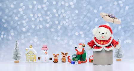 White teddy bear emerges from open gift box with row of Santa Claus dolls and many Christmas decorations on blurred snowing decorative  backgroundの写真素材