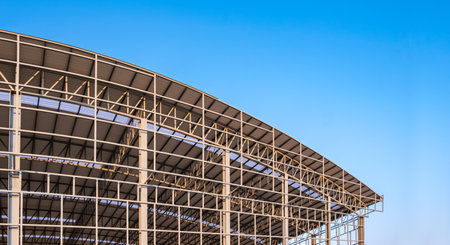 Part of large metal industrial factory building structure with corrugated steel curve roof and skylights in construction site against blue sky background, low angle viewの写真素材