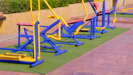 Row of colorful outdoor metal exercise equipment group on green turf and red cobblestone tile floor in public park areaの写真素材