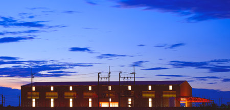 Silhouette industrial building structure with illuminated electric lighting inside against blue hour...