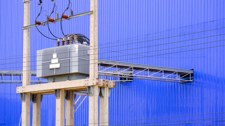 Electrical transformer and high voltage distribution equipment on electric pole with overhead power cable ladder tray next to blue aluminium corrugated metal wall of industrial facの写真素材