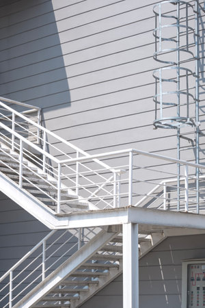 White metal fire escape with cylindrical ladder on gray industrial office building wall background, low angle view and vertical frameの写真素材