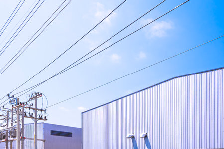 Two white factory warehouse industry buildings with high voltage electric power poles and cable lines against blue sky background, low angle and perspective side viewの写真素材